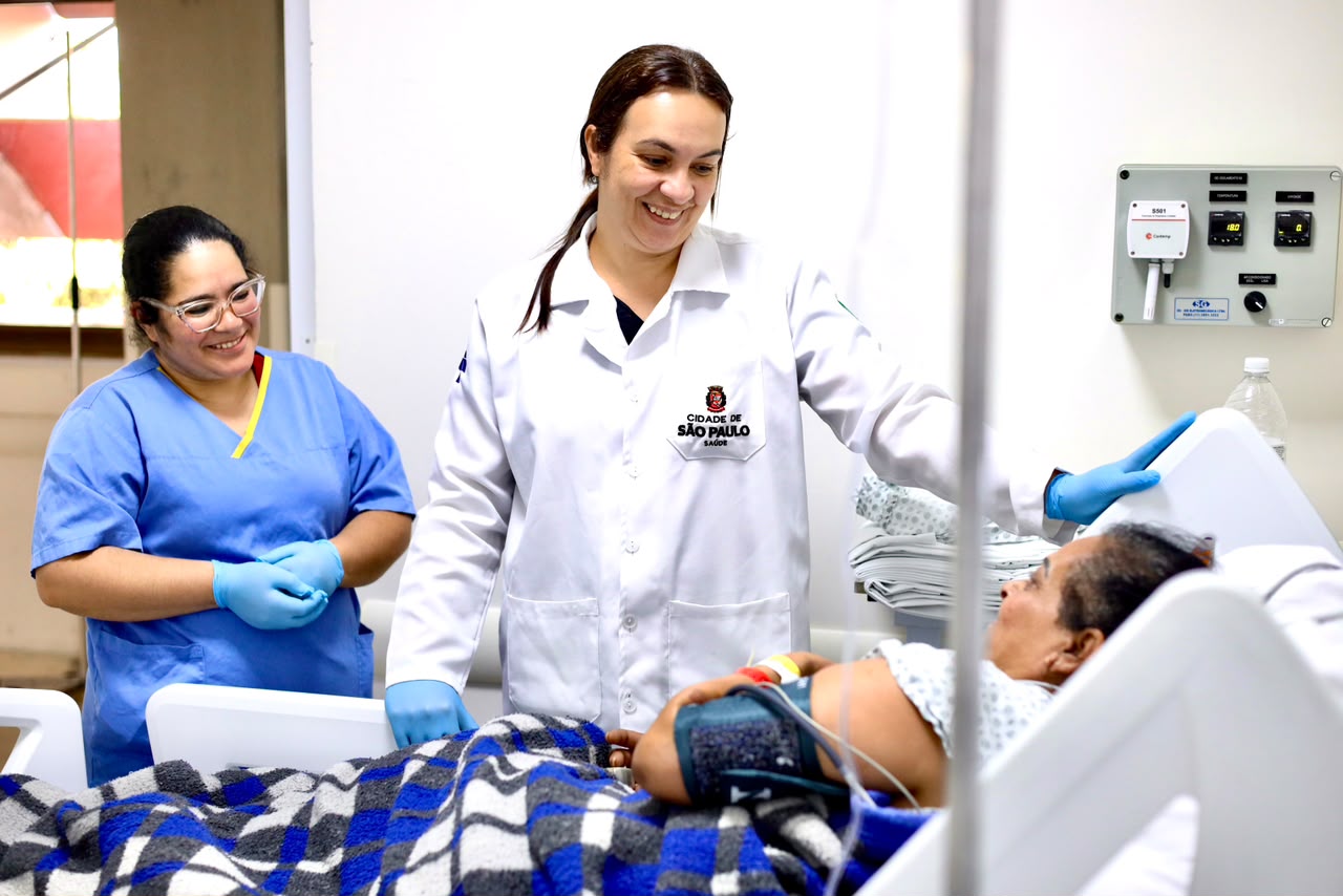 Duas profissionais de saúde atendem uma paciente deitada em um leito hospitalar. Uma delas, de jaleco branco e luvas azuis, sorri enquanto conversa com a paciente. Ao lado, outra profissional, de uniforme azul e também usando luvas, acompanha o atendimento sorrindo. A paciente está coberta por uma manta xadrez azul e cinza e utiliza equipamentos de monitoramento. O ambiente é claro e transmite uma interação acolhedora entre a equipe e a paciente.
