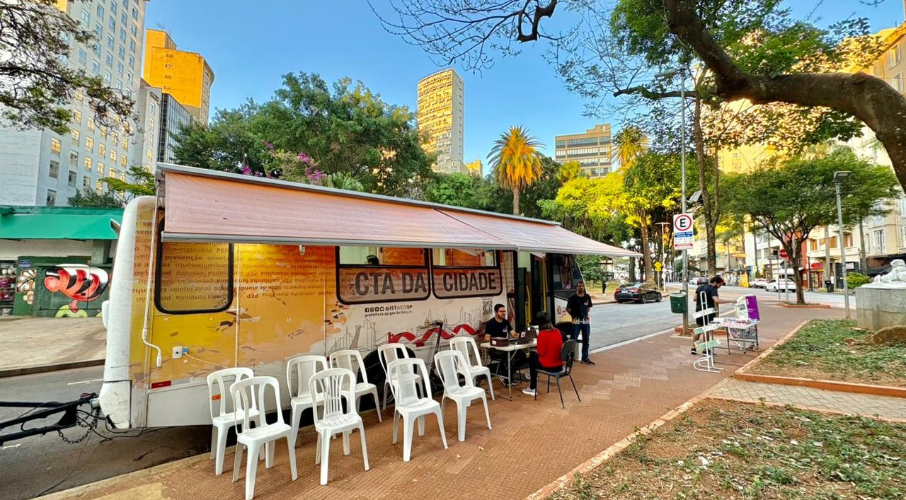 A imagem mostra uma unidade móvel de atendimento instalada em uma praça ou área pública arborizada de uma região central da cidade. Em destaque está um trailer adaptado, identificado como “CTA da Cidade”, sigla normalmente associada aos Centros de Testagem e Aconselhamento em saúde. O veículo possui uma cobertura retrátil aberta sobre a calçada, criando uma área de atendimento ao público. Em frente ao trailer há várias cadeiras plásticas brancas organizadas para espera, enquanto algumas pessoas conversam ou realizam atendimentos em mesas instaladas ao lado da unidade. O cenário urbano é composto por prédios altos ao fundo, árvores de grande porte, palmeiras e uma praça com áreas de circulação de pedestres. A luz do sol ilumina a cena, sugerindo que a atividade ocorre durante o dia.