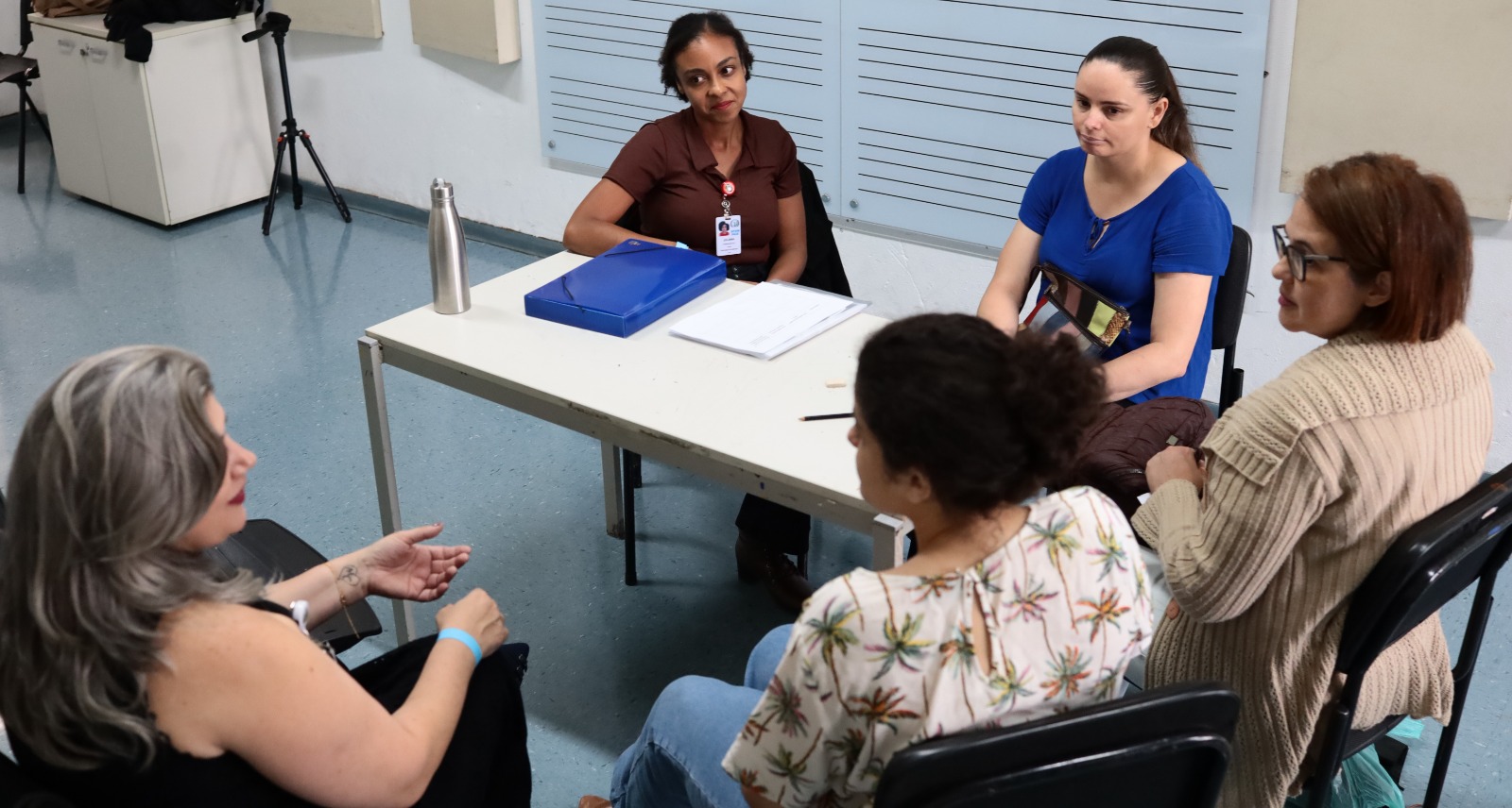 A imagem mostra um pequeno grupo de pessoas reunidas em uma sala que parece ser de um serviço público, possivelmente uma unidade de saúde ou espaço de atendimento.  Cinco mulheres estão sentadas ao redor de uma mesa simples. Duas delas, posicionadas atrás da mesa, parecem conduzir a conversa — uma usa crachá e tem uma pasta azul à frente, sugerindo que pode ser uma profissional de saúde ou facilitadora. As outras três mulheres estão sentadas do lado oposto, ouvindo com atenção; uma delas gesticula enquanto fala, indicando que está compartilhando alguma experiência ou fazendo uma pergunta.  Sobre a mesa há alguns materiais de trabalho, como uma pasta, papéis e uma garrafa térmica.