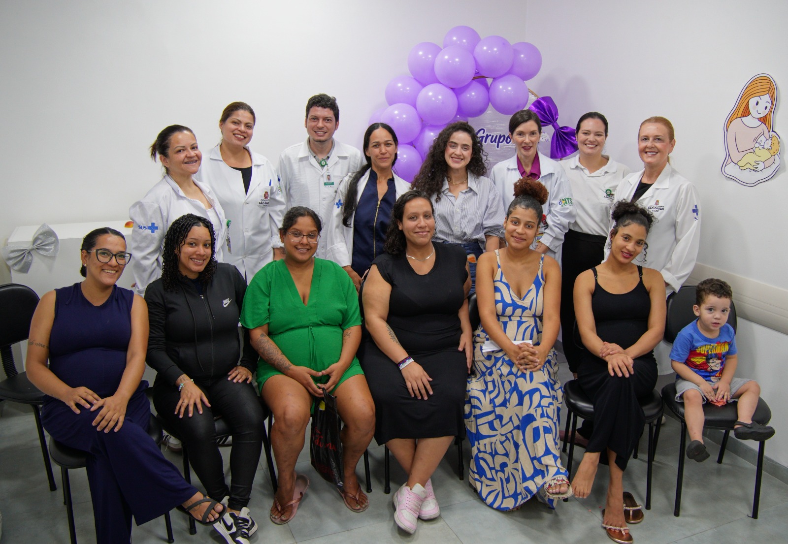 A imagem mostra um grupo reunido em uma sala clara da UBS Vila Constância, posando para uma foto coletiva. Na fileira da frente, há seis mulheres grávidas sentadas em cadeiras e uma criança pequena sentada à direita. As mulheres usam roupas casuais e variadas, incluindo vestidos, macacões e roupas esportivas, em cores como azul-marinho, preto, verde e estampado azul e branco. Atrás delas, há várias profissionais de saúde em pé, usando jalecos brancos, sorrindo para a câmera. O grupo parece diverso e acolhedor. Ao fundo, a decoração inclui um arco de balões lilás e roxos, além de um painel decorativo com temática materna e uma ilustração de uma mãe amamentando um bebê na parede. 