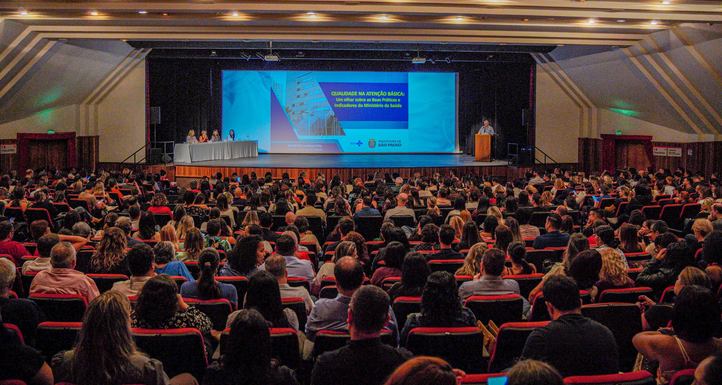 A imagem mostra um grande auditório lotado durante um evento institucional, com dezenas de pessoas sentadas em fileiras de poltronas vermelhas, todas voltadas para o palco.  No palco, há uma mesa com quatro participantes sentados à esquerda e um palestrante em pé à direita, falando em um púlpito. Ao fundo, um grande telão exibe uma apresentação com o título “Qualidade na Atenção Básica: um olhar sobre as boas práticas e indicadores do Ministério da Saúde”, além da identidade visual da Prefeitura de São Paulo.  O ambiente é bem iluminado e organizado, com estrutura de conferência, indicando um encontro técnico ou seminário voltado a profissionais da área da saúde, com foco em qualificação, indicadores e melhoria dos serviços públicos.