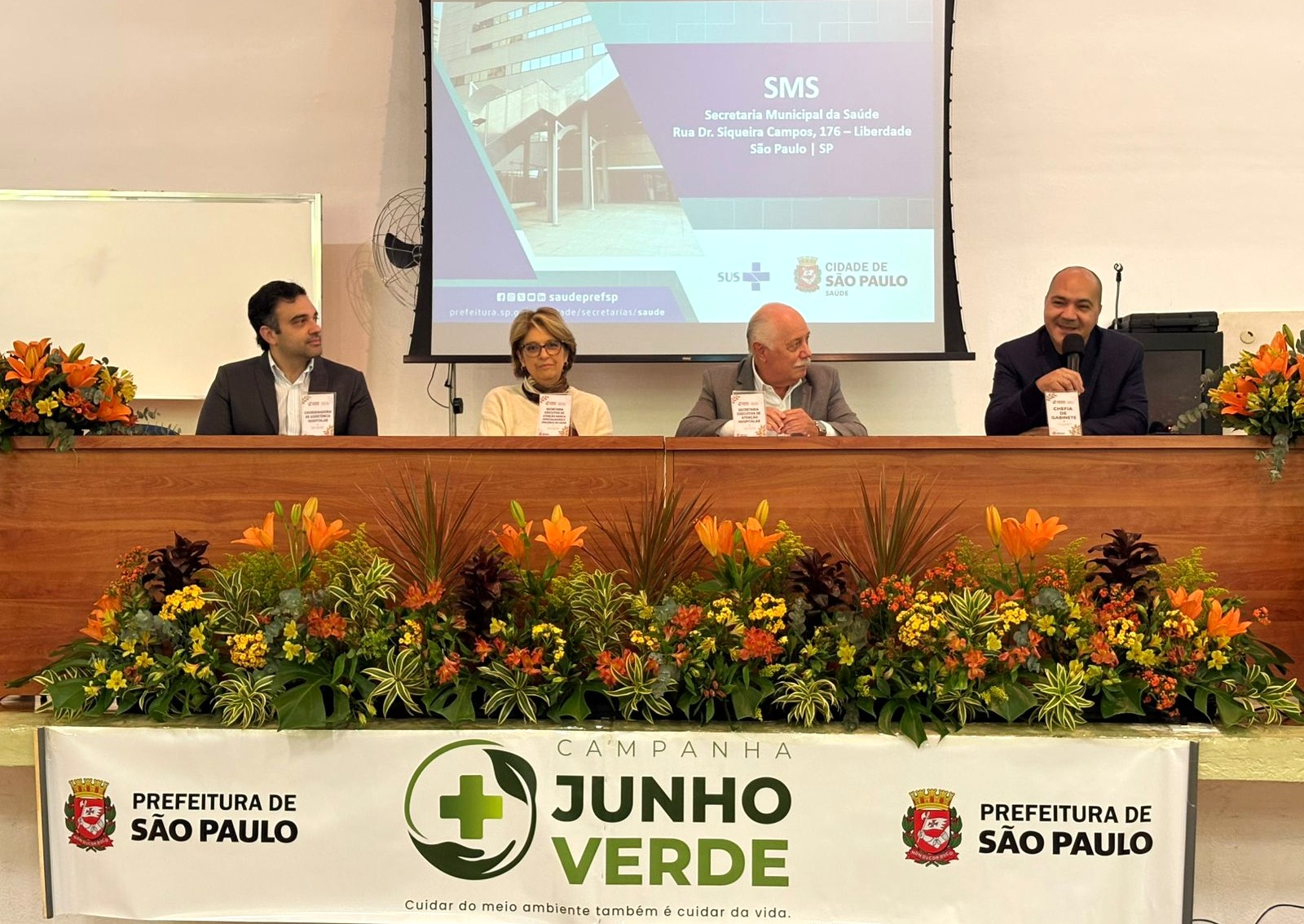 A imagem mostra uma mesa de autoridades durante um evento institucional realizado em auditório ou salão de conferências. Quatro pessoas estão sentadas atrás de uma longa bancada de madeira, ornamentada com um grande arranjo de flores e folhagens em tons de laranja, amarelo e verde.  Ao fundo, um telão exibe uma apresentação da Secretaria Municipal da Saúde (SMS) de São Paulo, com referência ao endereço da sede da pasta, na Rua Dr. Siqueira Campos, na Liberdade. Também aparecem os logotipos do SUS e da Prefeitura de São Paulo.  Na parte frontal da mesa há uma faixa da campanha “Junho Verde”, acompanhada do slogan “Cuidar do meio ambiente também é cuidar da vida”, além dos brasões da Prefeitura de São Paulo. O homem à direita está com um microfone em mãos e parece estar discursando ou participando de uma mesa-redonda, enquanto os demais acompanham a atividade.