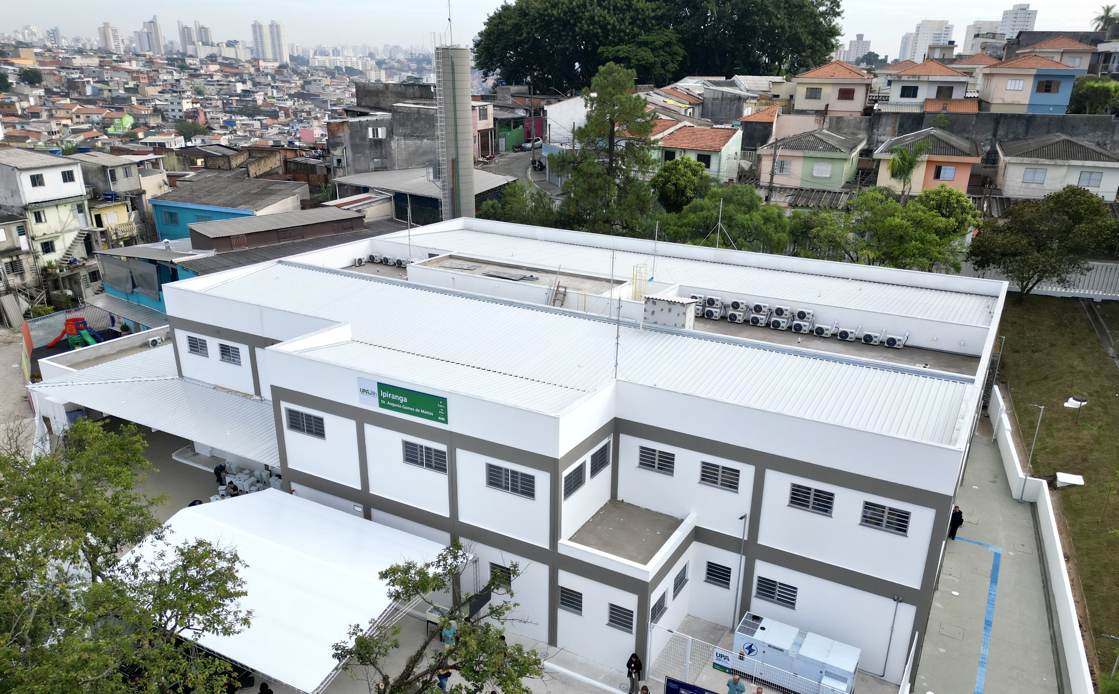 A imagem mostra uma vista aérea da Unidade de Pronto Atendimento (UPA) Ipiranga, que é um prédio grande, moderno e de cor clara. A estrutura tem formato retangular, com telhado metálico branco e várias janelas distribuídas pelas paredes externas. Na lateral do edifício, há uma cobertura branca onde algumas pessoas estão reunidas, possivelmente aguardando atendimento. Próximo a essa área, é possível ver uma pequena área com brinquedos coloridos, sugerindo um espaço infantil. No topo do prédio, há diversos equipamentos instalados, como aparelhos de ar-condicionado alinhados e uma estrutura vertical que parece ser um reservatório ou torre técnica. Ao redor do edifício, há árvores e áreas abertas. O entorno mostra um bairro bastante denso, com muitas casas pequenas e construções próximas umas das outras, além de prédios altos ao fundo, indicando uma área urbana extensa. Também há uma rua lateral com calçada ao lado do prédio.