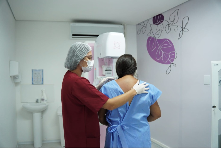 Fotografia em ambiente interno de uma sala de exames com paredes claras. À esquerda, uma profissional da saúde de perfil, com touca cirúrgica branca, máscara de proteção no rosto e jaleco ou uniforme na cor vermelha, posiciona uma paciente para um exame. Ela usa luvas brancas e apoia delicadamente as mãos nos ombros da paciente. A paciente está de costas, com os cabelos longos e soltos, vestindo um avental hospitalar azul claro amarrado nas costas. À frente delas, há um equipamento de mamografia branco e rosa. Ao fundo, aparecem uma pia, um ar-condicionado na parede e um adesivo decorativo com formas abstratas e tons de roxo na parede lateral.