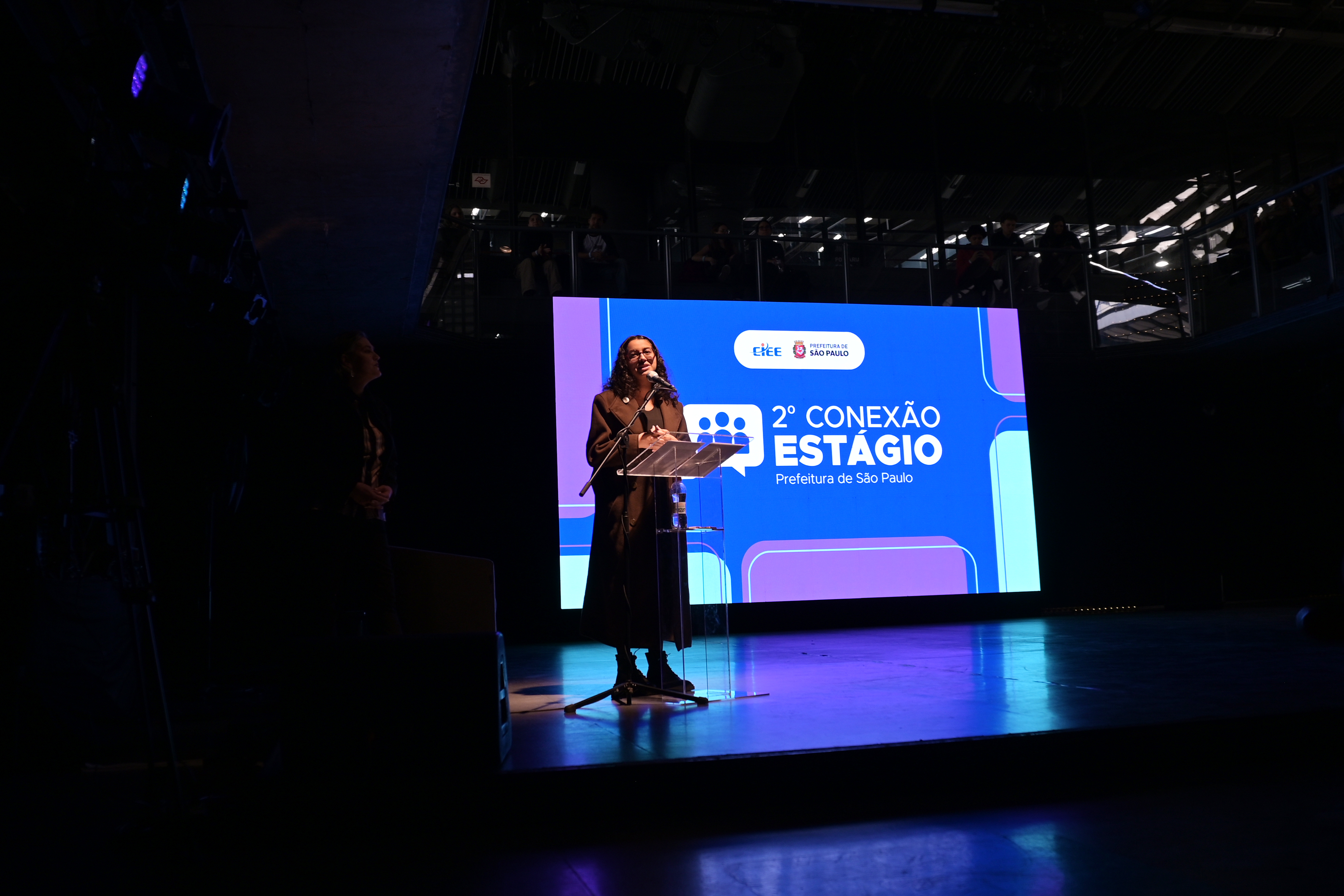 #ParaTodosVerem: Foto de uma mulher jovem de cabelos cacheados falando ao microfone em um púlpito transparente no palco. Atrás dela, há um grande telão azul com o texto
