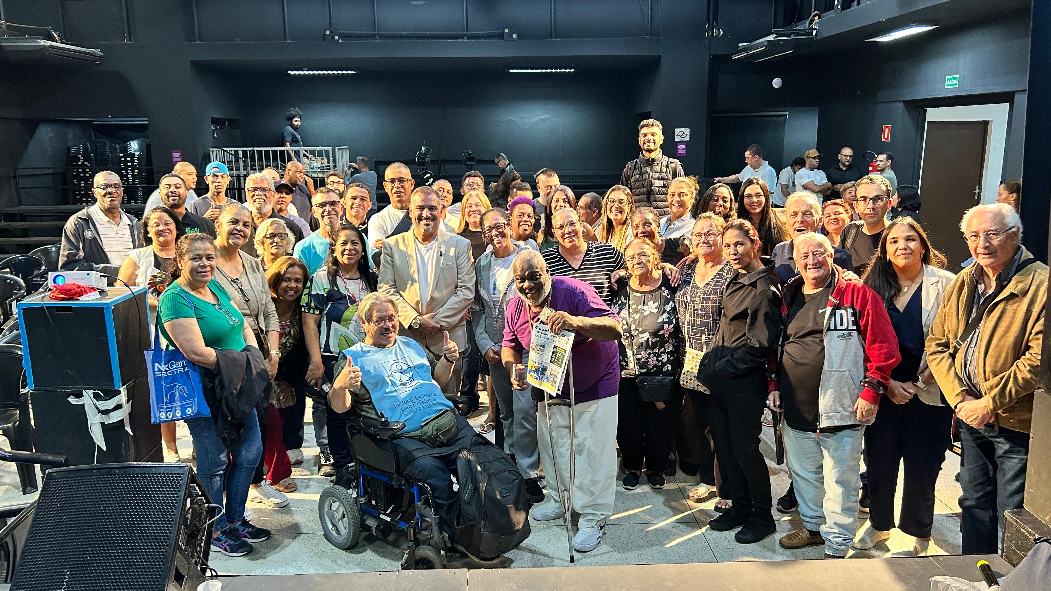Foto realizada após o evento A imagem mostra um grande grupo diverso de cerca de 50-60 pessoas reunidas em um espaço interno, como um auditório o. Elas posam para uma foto em grupo, com destaque para um homem em cadeira de rodas no centro à esquerda fazendo sinal de positivo com as mãos.