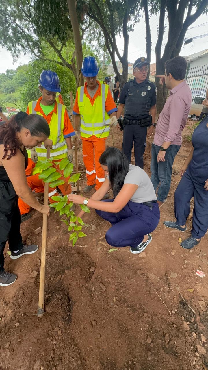 Duas Estudantes agachadas remexem na terra e uma delas usando enxadas realizam o plantio de muda. Ao lado dois zeladores usam uniformes na cor laranja, capacetes na cor azul, observam o plantio e estão ladeados pelo policial da GCM e pelo subprefeito da Cidade Tiradentes.
