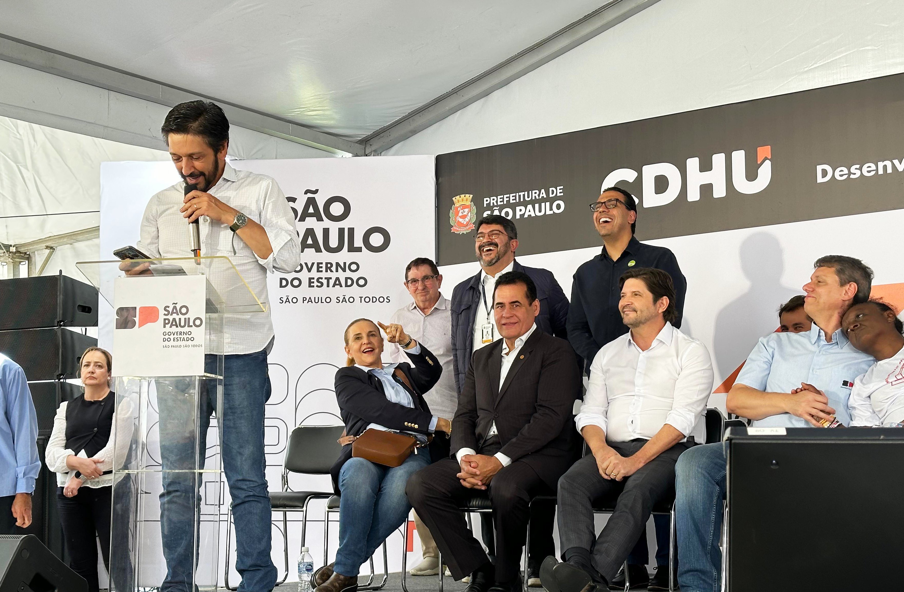 Em um palco coberto por uma tenda branca, várias pessoas participam de um evento oficial. À esquerda, o Prefeito de São Paulo, Ricardo Nunes, com camisa clara fala ao microfone em um púlpito transparente com o logotipo do Governo do Estado de São Paulo. Atrás e ao lado dele, outras pessoas estão sentadas e em pé, sorrindo e observando o discurso. No fundo, há painéis com os logotipos da Prefeitura de São Paulo e da CDHU (Companhia de Desenvolvimento Habitacional e Urbano). O clima é descontraído, com algumas pessoas rindo e interagindo entre si.
