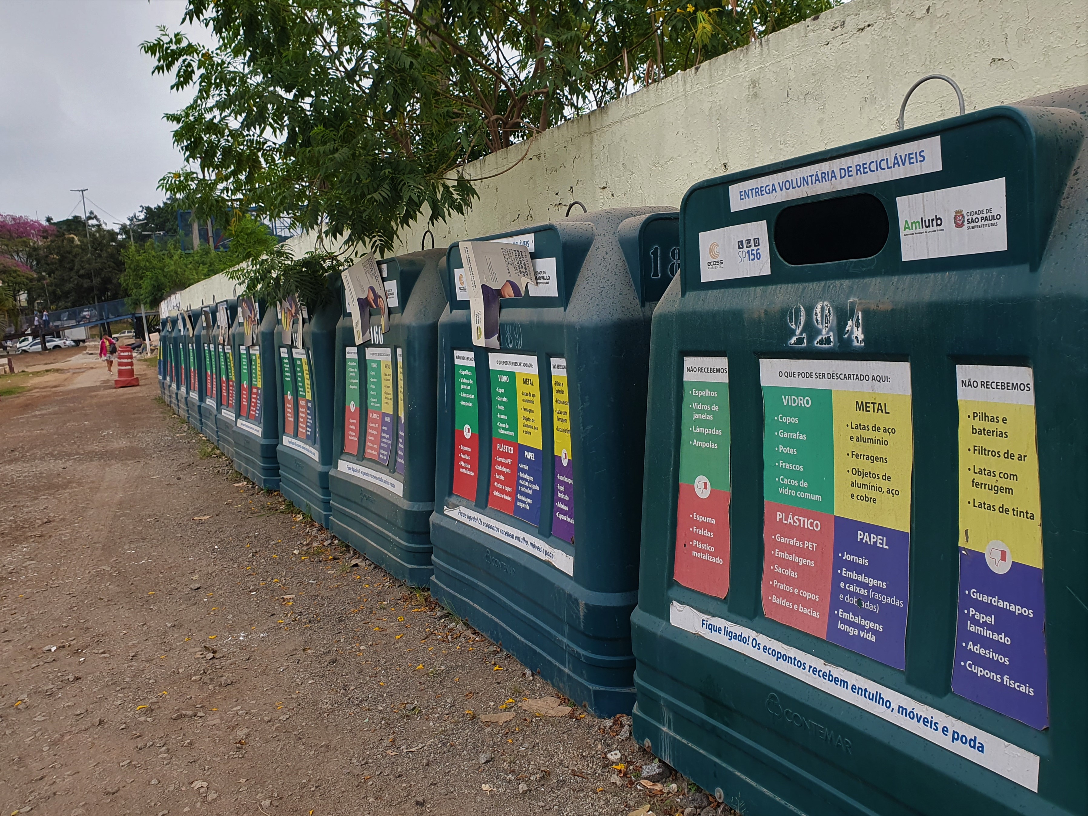 O registro foi feito em um ecoponto. Do lado direito da imagem até o fundo há lixeiras grandes de coleta seletivas em perspectiva. Todas são verdes. Em cada uma há a descrição dos diferentes tipos de material que podem ser descartados. Verde para vidro, Amarelo para metal, Vermelho para plástico e Azul para papel.