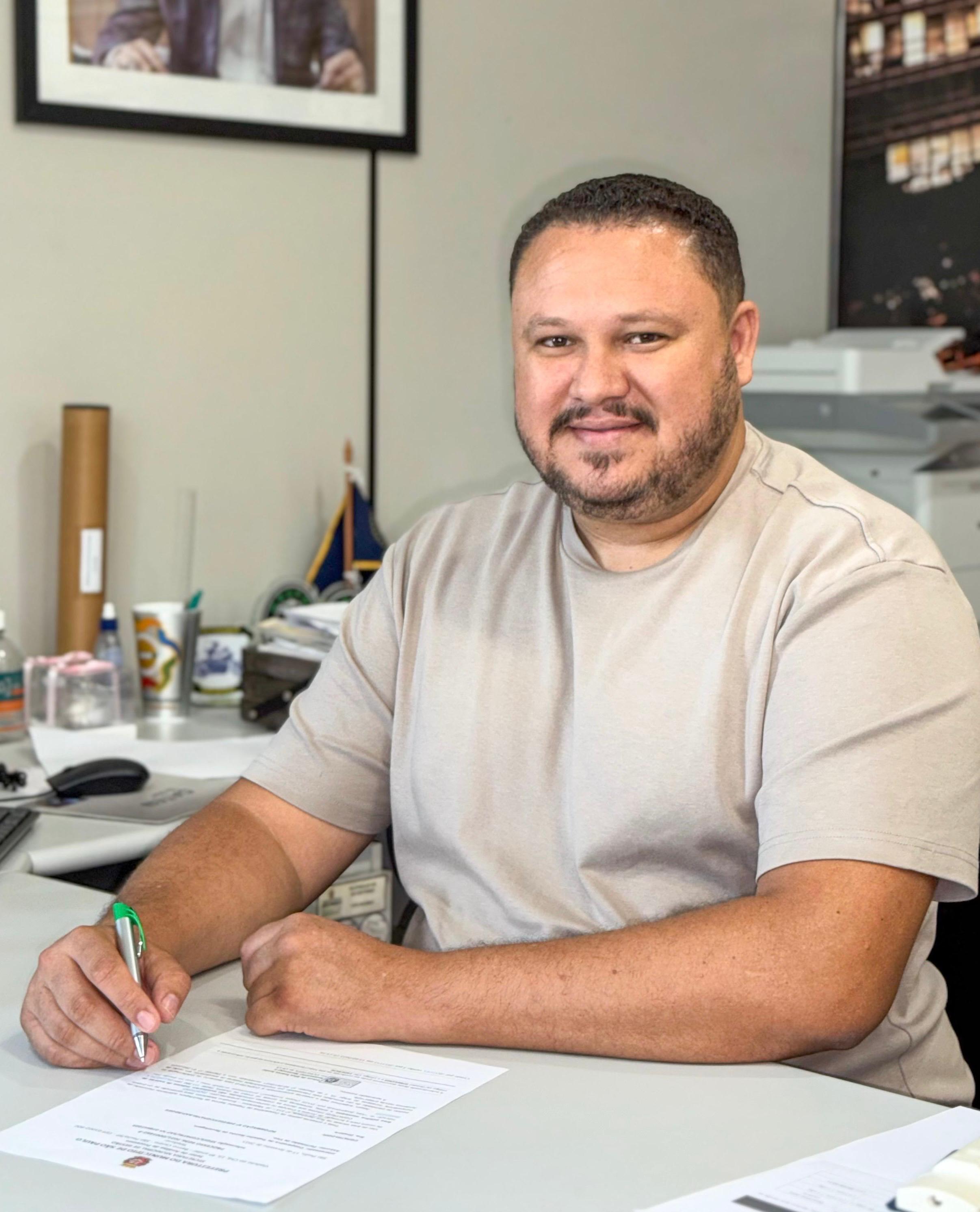 Um homem de pele clara, com cabelos curtos e escuros e barba rala escura, está sentado à mesa e sorri levemente para a câmera. Ele veste uma camiseta bege de mangas curtas. Sua mão direita está segurando uma caneta verde e branca sobre um documento que parece ser um formulário ou contrato, com selos ou logotipos visíveis na parte inferior. Sua mão esquerda repousa sobre a mesa ao lado do documento.  A mesa é clara e parece ser de cor cinza ou bege claro. Sobre ela, há outros objetos desfocados, incluindo o que parece ser um mouse de computador à esquerda, e vários copos ou canecas e papéis empilhados ao fundo. Atrás do homem, há uma parede clara e um quadro com uma imagem emoldurada na parte superior esquerda. À direita, parcialmente visível, parece haver uma estante ou prateleira com mais objetos. A iluminação parece vir da frente, iluminando o homem de forma suave.