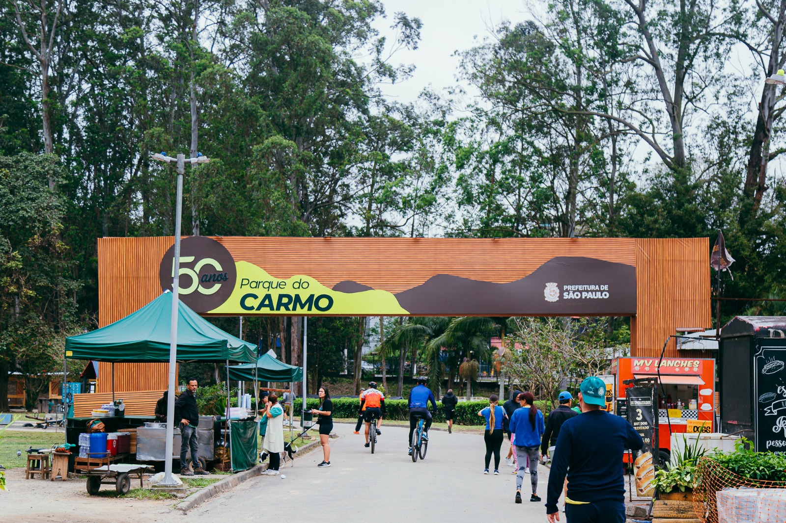 Descrição geral: Fotografia ao nível dos olhos, tirada ao ar livre durante o dia, mostrando a entrada do Parque do Carmo, situado em São Paulo.   Elemento central: Um grande pórtico horizontal em madeira com uma placa decorativa comemorativa dos 50 anos do Parque do Carmo, apresentando ainda o logotipo da Prefeitura de São Paulo no lado direito.   Primeiro plano: Uma estrada asfaltada que serve de caminho pelo parque, onde se encontram várias pessoas a passear a pé e ciclistas a andar de bicicleta.   Lados: À esquerda, existem tendas verdes de comércio ou apoio, carrinhos de comida de rua (food trucks) e caixas térmicas. À direita, há pequenas estruturas de comércio de lanches com letreiros e sinalética com os preços.   Fundo: Uma vegetação densa com árvores altas e copudas que preenchem todo o horizonte sob um céu claro e ligeiramente nublado