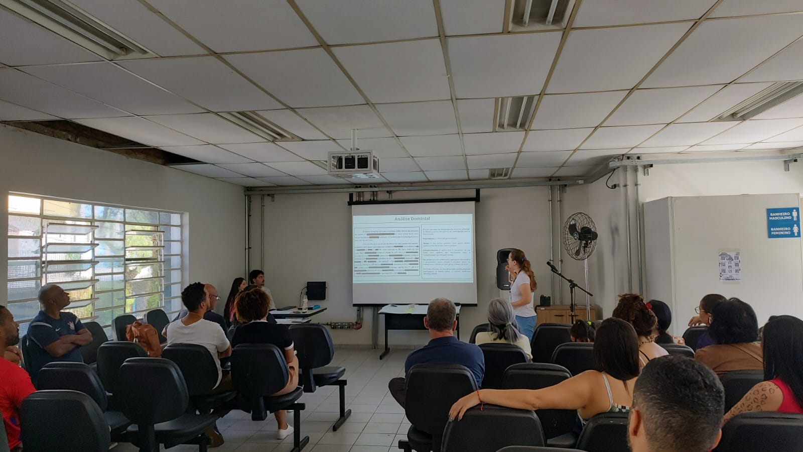 Sala de reunião com moradores sentados em cadeiras, assistindo a uma apresentação projetada na parede enquanto uma mulher fala à frente.