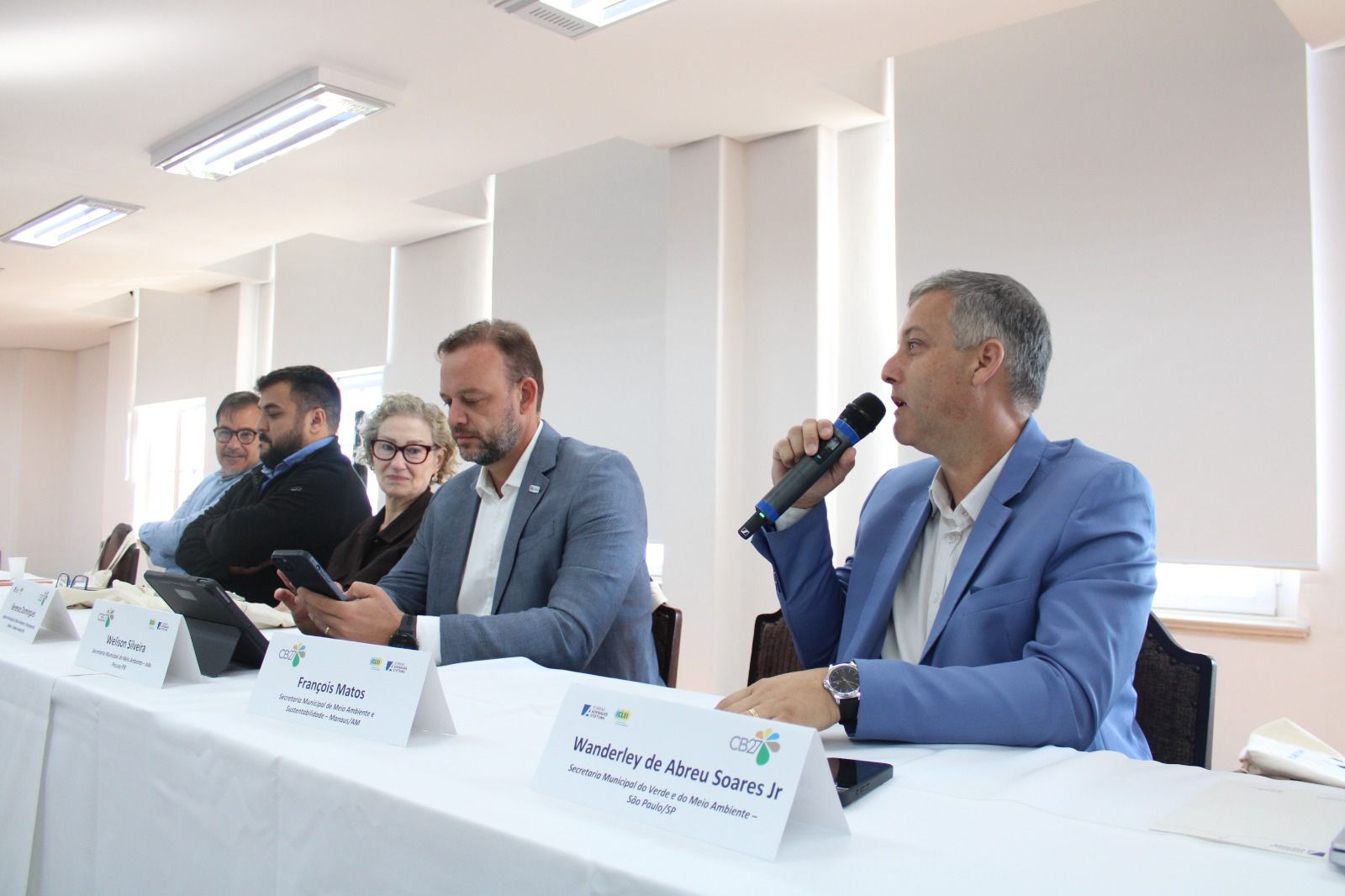 Homens e uma mulher estão sentados lado a lado em uma mesa comprida coberta por toalha branca, em uma sala bem iluminada. Eles participam de uma mesa de debate ou reunião formal. À direita, um homem de blazer azul segura um microfone e fala, voltado para o público. Ao lado dele, outro homem de blazer cinza olha para o celular. Mais à esquerda, uma mulher de óculos observa a cena, enquanto outros dois homens também acompanham a atividade. Sobre a mesa, há placas com nomes e cargos, além de celulares e tablets. O ambiente tem paredes claras, janelas com persianas e iluminação de teto.