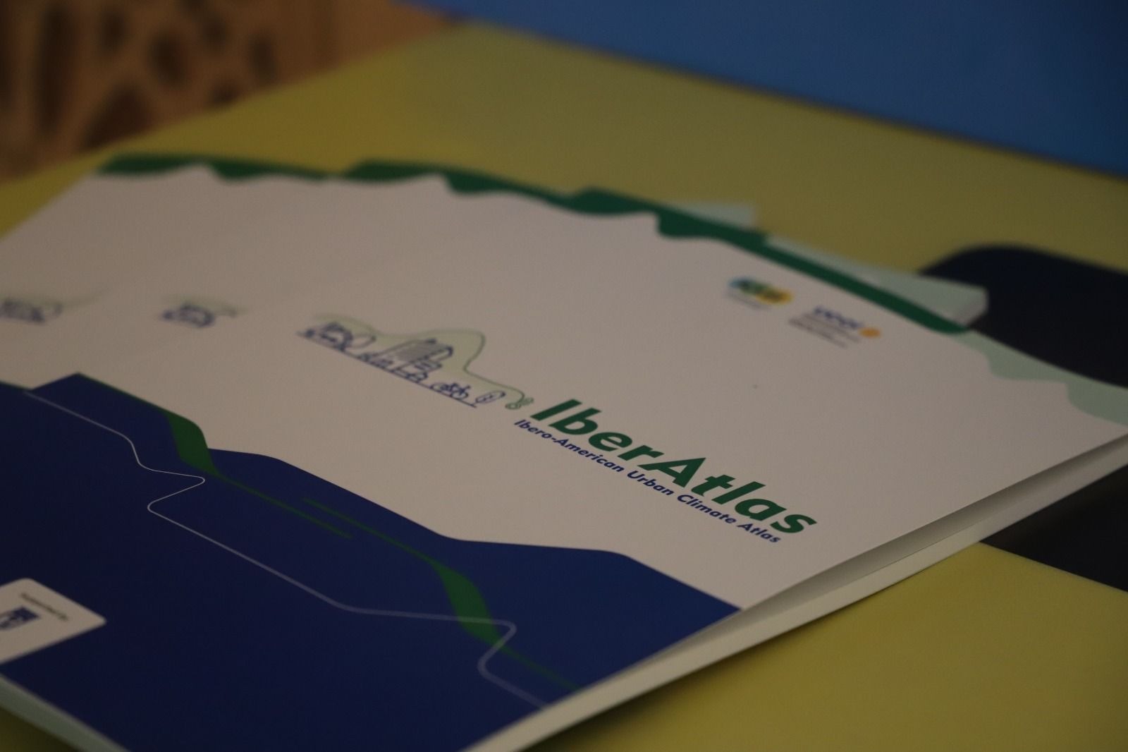 Fotografia em close de um conjunto de cadernos ou pastas empilhados sobre uma mesa. Em primeiro plano, aparece uma capa branca com elementos gráficos em verde e azul e o título "IberAtlas – Ibero-American Urban Climate Atlas". Ao fundo, outras capas semelhantes estão parcialmente sobrepostas e desfocadas. A iluminação é suave e o foco destaca a publicação, enquanto o restante da cena permanece com profundidade de campo reduzida.