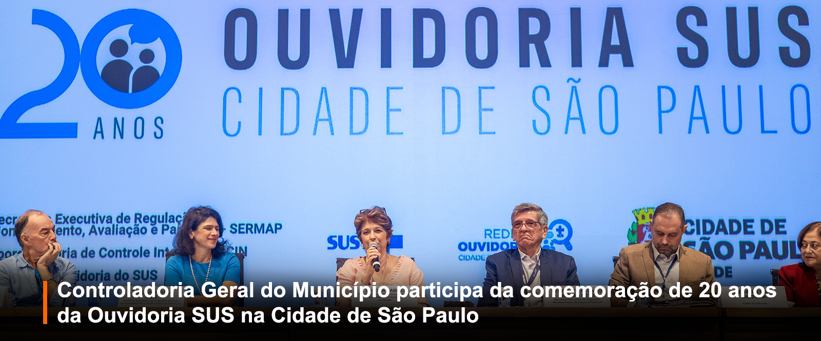 Fotografia de evento formal em auditório, com seis autoridades em mesa. Painel azul ao fundo com a logomarca dos 20 anos da Ouvidoria SUS da Cidade de São Paulo. Faixa preta inferior: 
