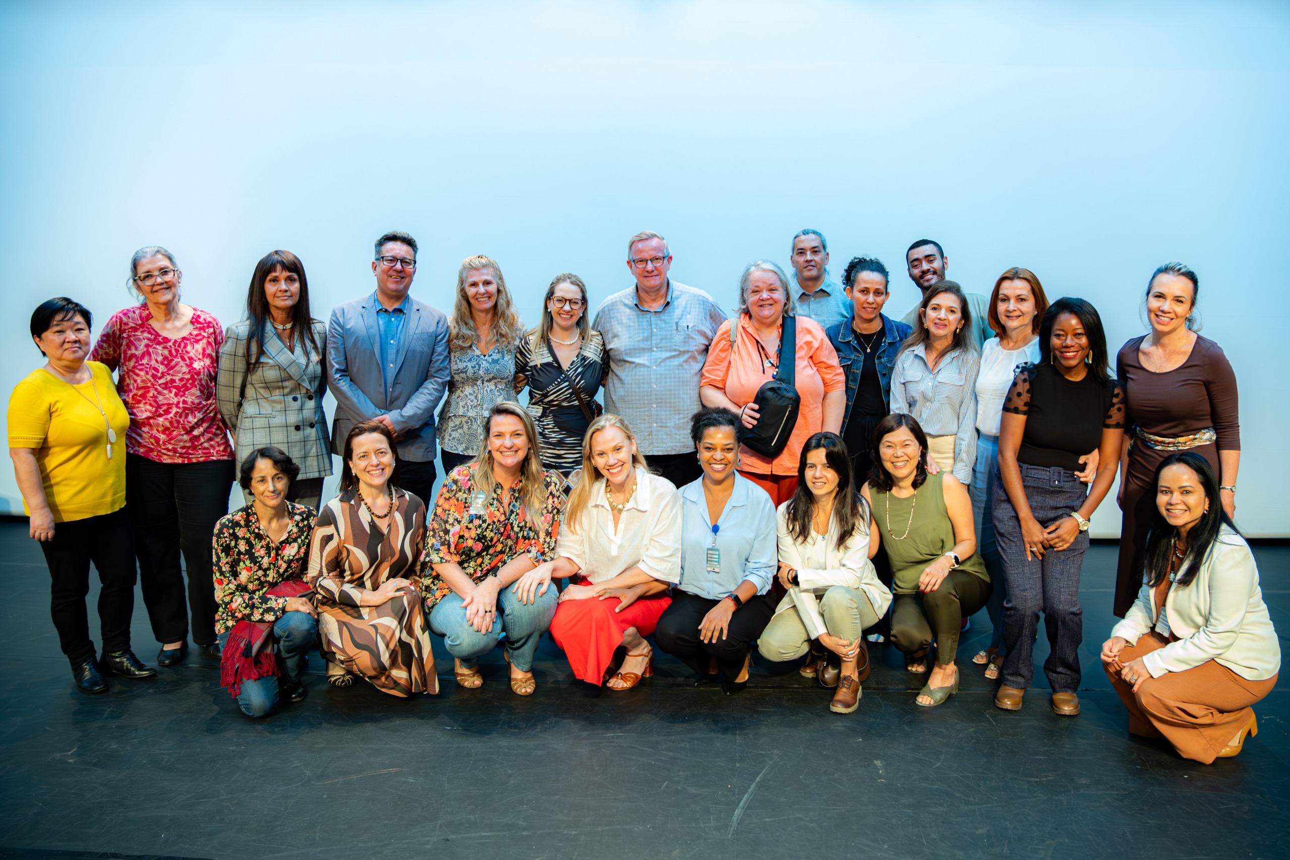 A imagem mostra um grupo grande de pessoas reunidas para uma foto coletiva em um ambiente interno, provavelmente um auditório ou palco. Ao todo, cerca de duas dezenas de participantes aparecem organizados em duas fileiras: uma em pé ao fundo e outra agachada ou sentada à frente.  O grupo é diverso em idade, gênero e aparência, com pessoas vestindo desde roupas mais formais, como blazers e camisas sociais, até trajes mais casuais. Muitos estão sorrindo e próximos uns dos outros, sugerindo um clima de integração, equipe ou encerramento de evento.  O fundo é neutro, com uma parede clara e iluminação uniforme, o que reforça o foco nas pessoas. A composição indica que se trata possivelmente de uma equipe de trabalho, participantes de um projeto ou integrantes de um evento institucional ou formativo.
