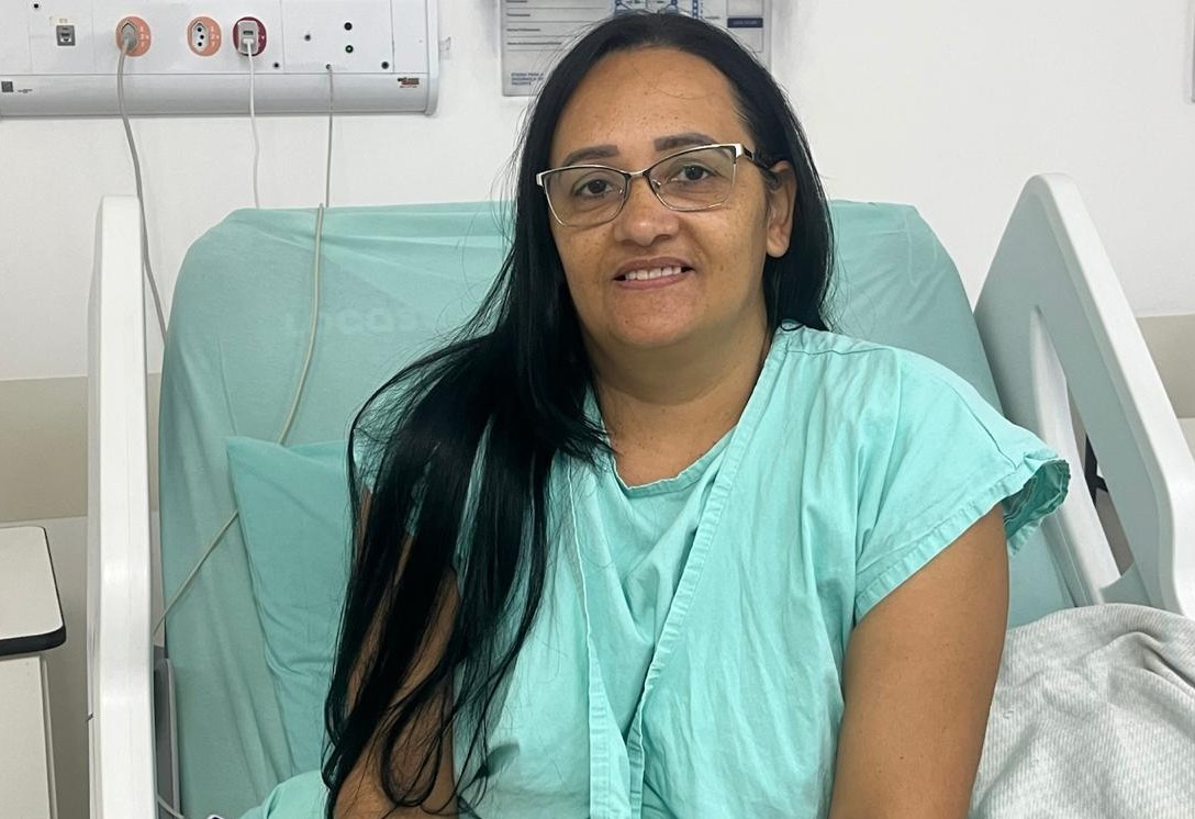 A imagem mostra uma mulher sentada em uma cama hospitalar, em um quarto de internação. Ela veste uma camisola hospitalar verde-clara e usa óculos de armação fina. Está posicionada de frente para a câmera, com expressão serena e um leve sorriso.  Ao fundo, é possível ver parte da estrutura do leito e equipamentos hospitalares instalados na parede, como pontos de gases medicinais e conexões utilizadas em ambientes assistenciais. A iluminação é clara e o ambiente aparenta estar limpo e organizado