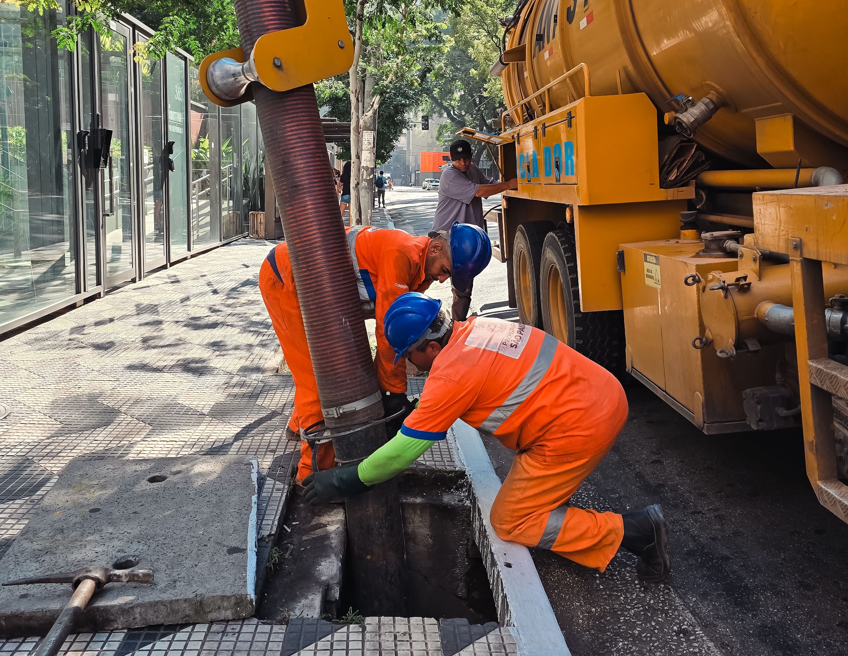 Fotografia em plano médio mostra três trabalhadores realizando um serviço de manutenção urbana na calçada. À direita, está estacionada a lateral de um grande caminhão amarelo de hidrojato.  No centro da imagem, dois trabalhadores aparecem curvados, operando uma grande mangueira de sucção grossa e sanfonada, que está inserida diretamente dentro de um bueiro aberto no chão. Um terceiro trabalhador está ao fundo, próximo aos comandos na lateral do caminhão.  A calçada é de blocos cinzas e, em primeiro plano, à esquerda, há uma tampa de concreto removida e uma ferramenta de ferro apoiada no chão. Ao fundo, vê-se uma estrutura de vidro e metal e uma rua arborizada.