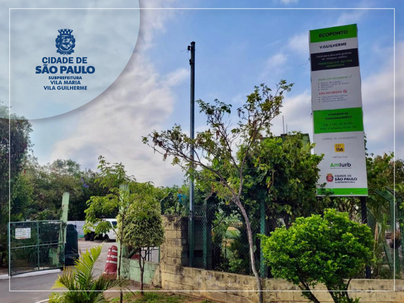 A imagem é uma fotografia externa que captura a entrada de um local arborizado, possivelmente uma área de serviço ou um centro comunitário, sob um céu parcialmente nublado.  No canto superior esquerdo, há uma marca d'água circular com o logotipo da Cidade de São Paulo (o brasão), e o texto que o acompanha diz: "CIDADE DE SÃO PAULO" e, em letras menores, "SUBPREFEITURA VILA MARIA / VILA GUILHERME".